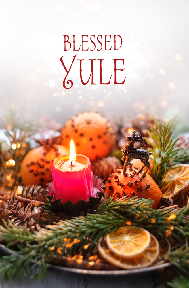 Red candle burns in center of Yule centerpiece with oranges, closed, pine branches, and the words Blessed Yule above.
