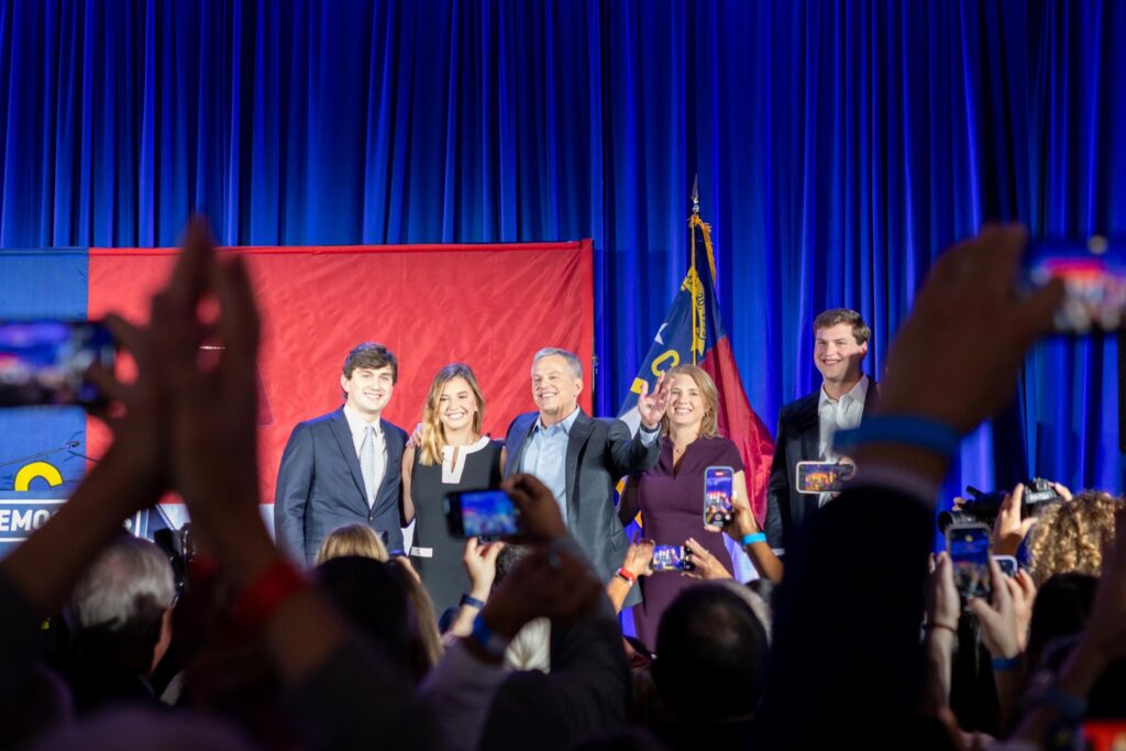 New North Carolina Governor Josh Stein on platform surrounded by family members celebrating November 2024 victory. People in foreground snapping photos.