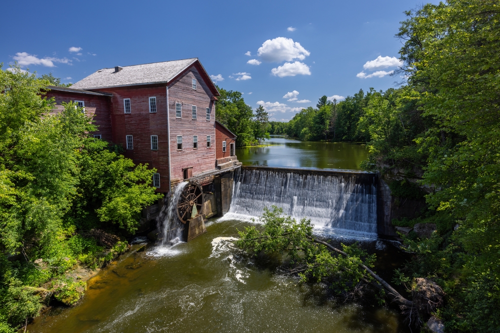 Old red wooden building next to stream with waterwheel and dam, surrounded by green trees and blue sky
